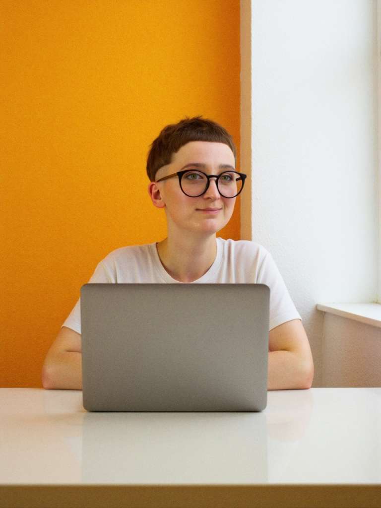 Person mit kurzem braunen Haar und dunkler Brille sitzt an einem Tisch und arbeitet an einem grauen Laptop. Die Wand im Hintergrund ist orange, das Fenster rechts bringt Tageslicht ins Bild.